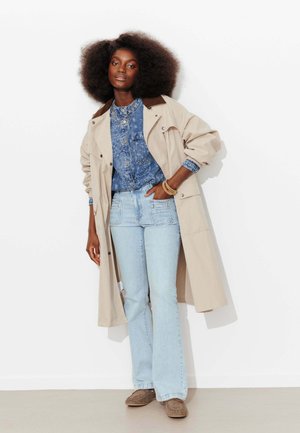 Femme avec une coiffure afro portant un trench-coat beige, une chemise en denim bleu à motifs, un jean bleu clair et des mocassins marron, debout contre un mur blanc.