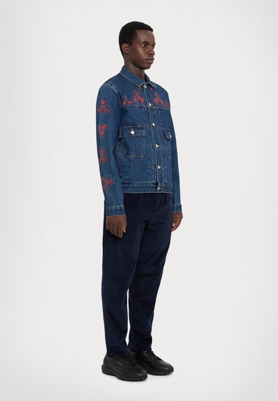 Denim jacket with red floral embroidery, two chest pockets, and a button-up front; paired with navy pants and black sneakers.