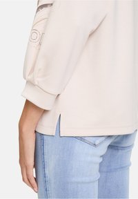 Pull léger beige avec des manches bouffantes, des fentes sur les côtés et un imprimé texturé. Associé à un jean bleu pâle, présentant un fini denim lisse.