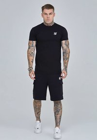 Sort T-shirt med korte ærmer og logo på brystet, kombineret med sorte cargoshorts. Begge lavet af stretchstof. Hvide sneakers fuldender looket.