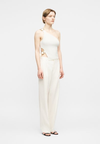 Γυναίκα που φοράει λευκό τοπ με έναν ώμο και λεπτομέρειες με χρυσούς κρίκους, μαζί με λευκό παντελόνι ψηλόμεσο, με φαρδιά μπατζάκια και μαύρα σανδάλια ανοιχτά στο μπροστινό μέρος.