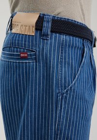 Mėlynos dryžuotos džinsai su juodai numečiamu diržu, kuriuos puošia odinė etikete su užrašu "BIG STAR" ir šoniniu kišenių detaliu.