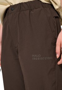 Bruine, lichte broek met een elastische tailleband, zijzakken en de bedrukte tekst "HALO 196819721981" op de voorkant. Gladde textuur.