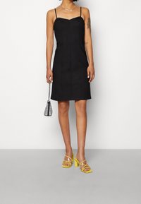 Robe noire ajustée avec des bretelles fines, présentant un décolleté droit et des coutures discrètes. Associée à des sandales à talons jaunes et à un petit sac à main.