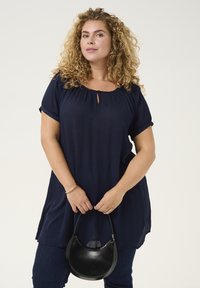 Blusa blu navy con maniche corte, scollo arricciato e dettaglio a chiave. Modella che tiene una piccola borsa nera. Jeans in denim.