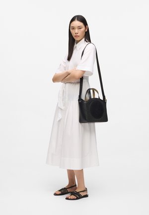 Ženska v beli midi obleki in črnih sandalah stoji z rokami prekrižanimi, s črno torbico z dolgo vrvico čez ramo.