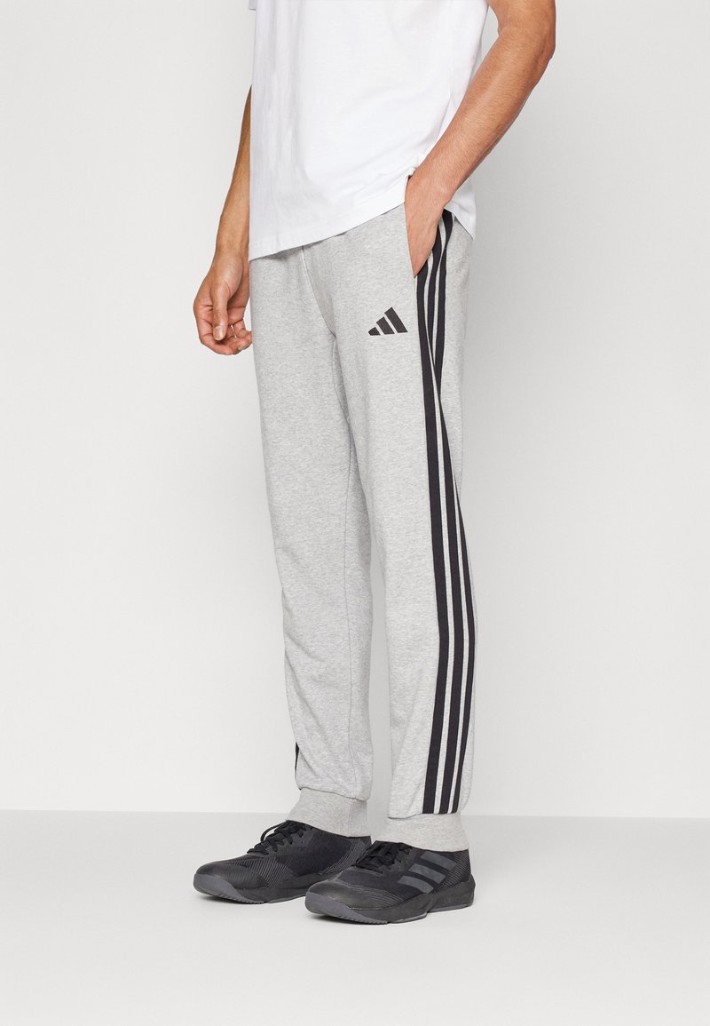Grå joggebukser med svart tre-stripes design og logo på venstre lår, med en avslappet passform og mansjetter på anklene.
