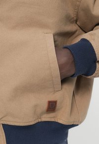 Hnedá plátenná bunda s námorníckymi modrými rebrovanými manžetami a bočným vreckom, s hnedou koženou logovou nášivkou v spodnej ľavej časti. Odolná textúra.