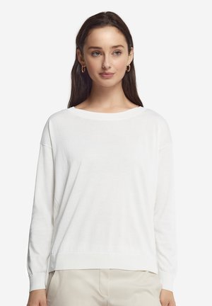 Kvinde med langt mørkt hår iført en hvid langærmet sweater og beige bukser, stående mod en simpel lys baggrund.