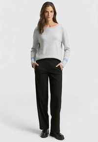 Pull gris clair texturé avec un col rond et des accents bleus sur les poignets, associé à un pantalon noir à jambes larges et des chaussures noires.
