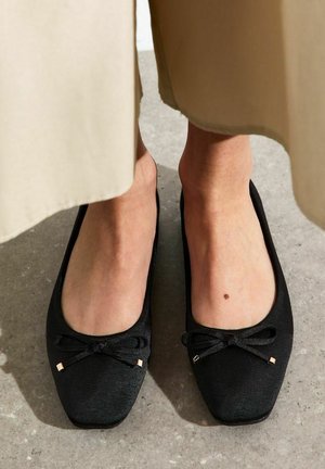 Feet wearing black square-toe ballet flats with bows, standing on a gray concrete floor, beige skirt hem visible above ankles.