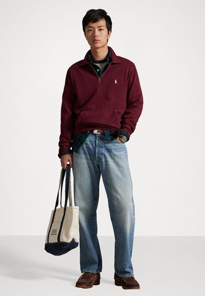 Bordeaux ritsfleece, blauw geruit overhemd eronder, lichtblauwe jeans en bruine schoenen. Canvas schoudertas met marineblauwe accenten.