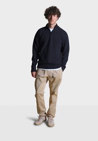 Námořnický svetr na zip, volné béžové kalhoty s nastavitelným pásem a světle barevné boty s akcenty. Jednoduché pozadí.