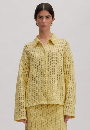 Frau mit dunklem Haar trägt gelben offenen Strick-Cardigan mit Knöpfen und passenden Rock, stehend vor hellem, einfachem Hintergrund.