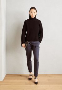 Pulover negru cu guler înalt din tricot cu model texturat, pantaloni slim fit bleumarin și pantofi negri cu vârf ascuțit și accente circulare din aur, stând pe podea din lemn.