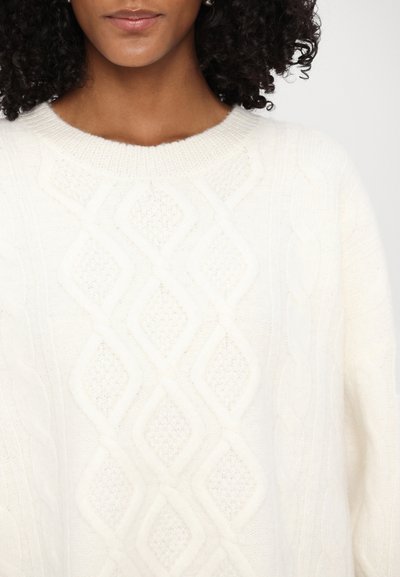Pull en tricot torsadé crème avec un motif en losanges, col rond côtelé et texture douce. Le tissu semble chaud et confortable.