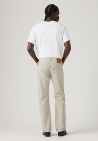 Man with braided hair wearing a white t-shirt, beige jeans, and black shoes standing with hands in back pockets facing away.