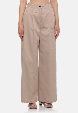 Pantaloni beige a gamba larga con pince frontali, chiusura con bottone, indossati con sandali bianchi con tacco e cinturini su una persona in piedi.