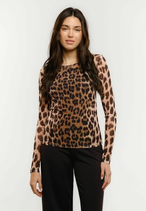 Jeune femme aux longs cheveux foncés portant un haut à manches longues imprimé léopard marron et noir et un pantalon noir, debout devant un fond uni.