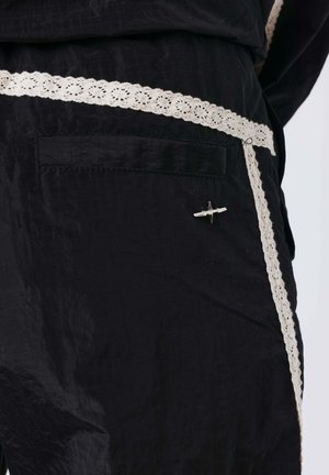 Zwarte broek met witte kanten zoom op de zak en zijnaden, met een klein zilveren vliegtuigspeldje op het zakgebied.