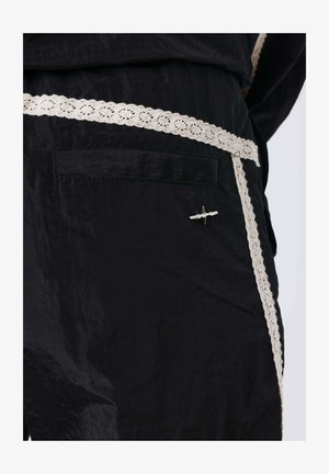 Zwarte broek met witte kanten zoom op de zak en zijnaden, met een klein zilveren vliegtuigspeldje op het zakgebied.