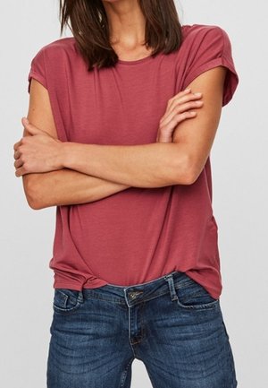 Femme portant une chemise rose poudré à manches courtes et un jean bleu, les bras croisés sur la poitrine devant un fond uni.
