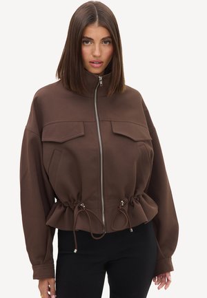 Femme aux cheveux bruns et lisses portant une veste marron zippée avec poches avant et taille ajustable par cordon, associée à un pantalon noir.