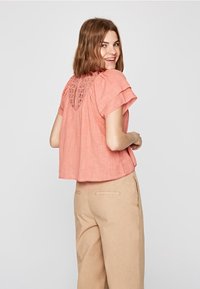 Blusa rosa chiaro a maniche corte con una vestibilità ampia, caratterizzata da un dettaglio in crochet sulla schiena e un tessuto testurizzato. Abbinata a pantaloni beige.