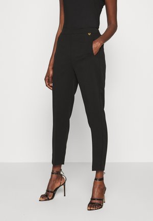 Kobieta ubrana w czarne spodnie zwężane, czarny bezrękawnik i czarne sandały na wysokim obcasie z paskami, stojąca z jedną ręką w kieszeni.