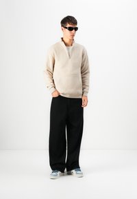 Beige fleece hoodie med en kvart lynlås og frontlomme, parret med løse sorte bukser og sneakers med blå accenter. Sorte solbriller.
