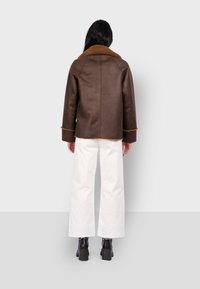 Brązowa skórzana kurtka z dużym jasnobrązowym kołnierzem, widocznym przeszyciem i szerokimi rękawami. Noszona z białymi spodniami o szerokich nogawkach i czarnymi masywnymi butami.