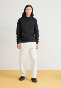 Zwarte katoen hoodie met een trekkoord, witte relaxte broek en zwarte sneakers. De achtergrond is lichtgrijs met een gele vloer.