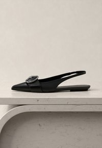 Μαύρο σανδάλι με αιχμητή μύτη και ασημένια στρογγυλή αγκράφα, εκτεθειμένο σε ένα φωτεινό πέτρινο τραπέζι.