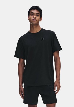 Jeune homme aux cheveux courts et bouclés portant un t-shirt noir et un short noir, debout devant un fond clair uni.