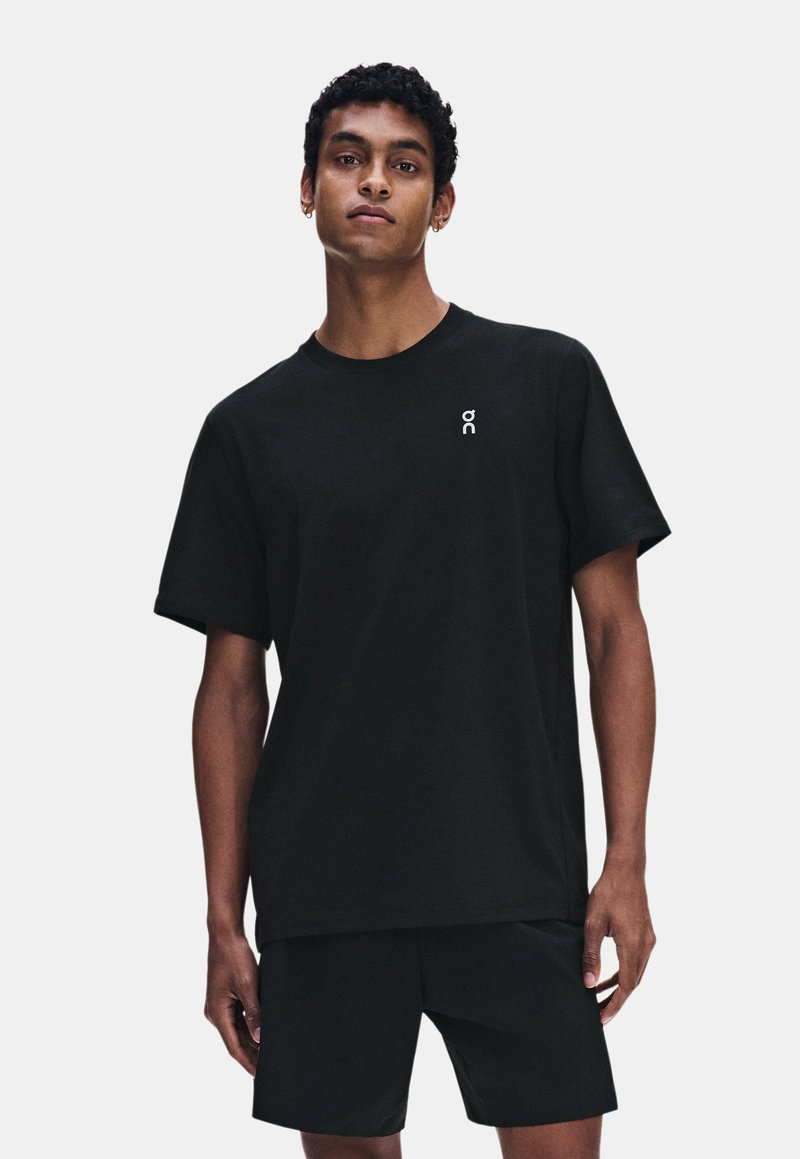 Jeune homme aux cheveux courts et bouclés portant un t-shirt noir et un short noir, debout devant un fond clair uni.