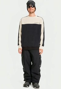 Schwarzer und beiger Farbblock-Sweatshirt mit langen Ärmeln, kombiniert mit schwarzen Cargo-Hosen. Abgerundet mit Logodetails und einer Mütze.