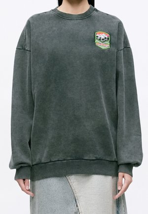 Personne portant un sweat-shirt vert délavé oversize avec des poignets et un ourlet côtelés, présentant un patch coloré sur la poitrine gauche.