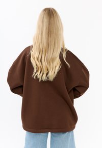 Brązowy oversize'owy sweter z opadającymi ramionami, charakteryzujący się miękką teksturą. W połączeniu z jasnoniebieskimi dżinsami o luźnym kroju, widoczny z tyłu.