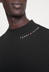 Man i svart högkrage-tröja med vit "Tommy Hilfiger" text och logotyp nära kragen på en neutral bakgrund.