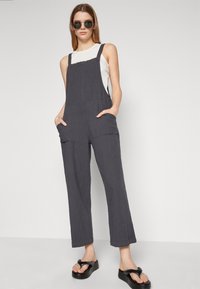 Dark gray fabric overall with wide legs, side pockets, and adjustable straps. Worn over a white top, paired with black platform sandals.