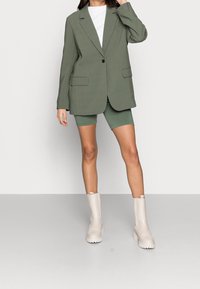 Blazer vert olive à coupe cintrée, avec revers en encoche, poches avant ; associé à un short assorti et des bottines beiges à semelle épaisse.
