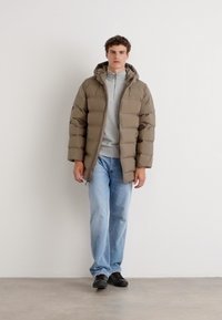 Beige pufferjack, met een capuchon en een gevoerd ontwerp. Draagt over een grijze trui, gecombineerd met lichtblauwe jeans en zwarte schoenen.