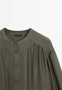 Blouse boutonnée vert olive foncé avec col rond et coutures froncées aux épaules, en tissu léger.