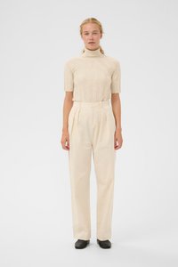 Kvinde med blont hår iført en beige ribbet turtleneck kortærmet bluse, højtaljede cremefarvede vide bukser og sorte sko, stående lige.