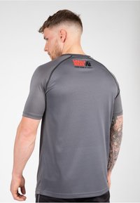 Camiseta deportiva de manga corta gris con un logo negro y rojo en la parte de atrás, con un diseño ajustado y textura suave.
