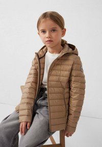 Tan pufferjas met horizontaal doorgestikt patroon, capuchon, ritssluiting en logo op de linkermouw, gedragen over een wit shirt en grijze broek.