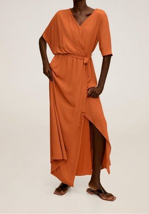 Femme portant une robe portefeuille fluide orange avec des manches aux coudes, une taille nouée, une fente haute et des sandales plates noires sur un fond uni.