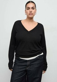 Čierny pulóver s výstrihom do "V", dlhými rukávmi a voľným strihom, skombinovaný s tmavými rifľami s kontrastným bielym pásom v páse.