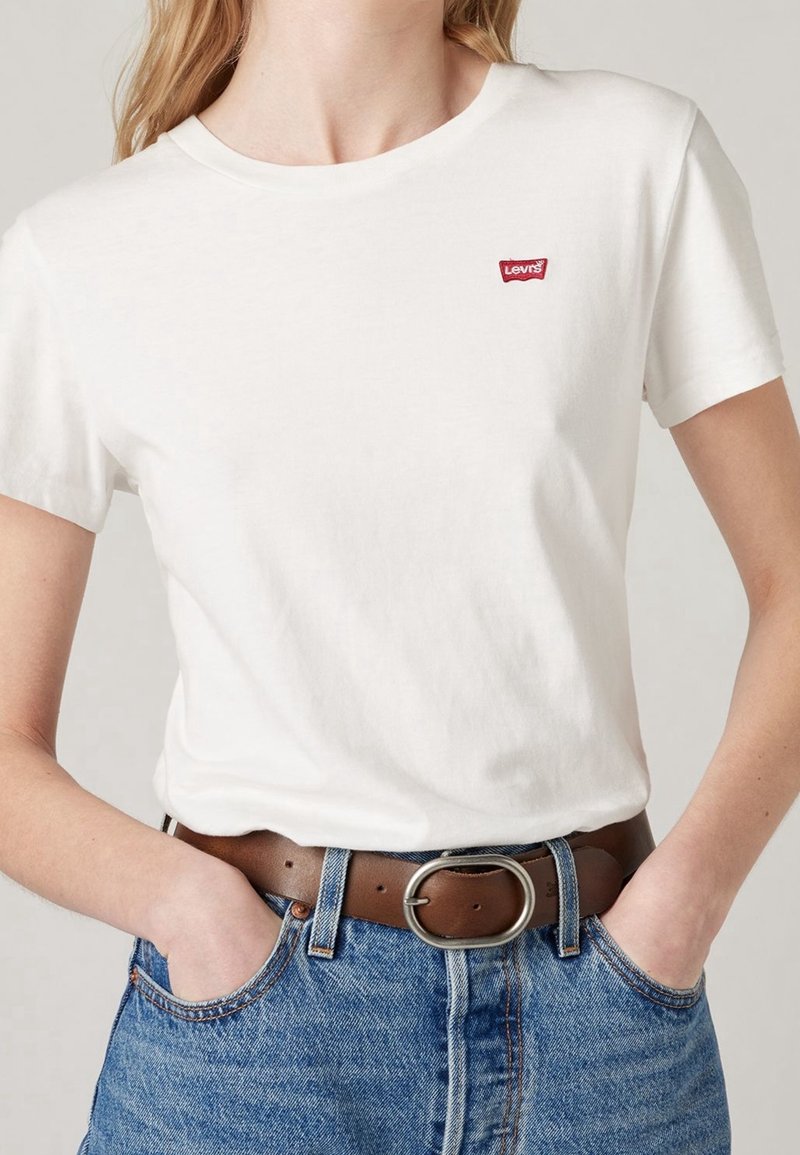 Femme portant un t-shirt blanc Levi's rentré dans un jean bleu avec une ceinture en cuir marron, mains partiellement dans les poches, montrée du cou aux hanches.