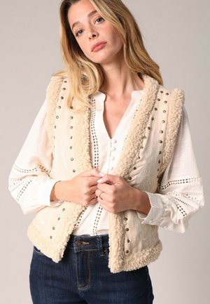 Beige mouwloos vest met pluizige schapenvachtafwerking, decoratieve studs en geborduurde details, gedragen over een witte blouse en donkere spijkerbroeken.